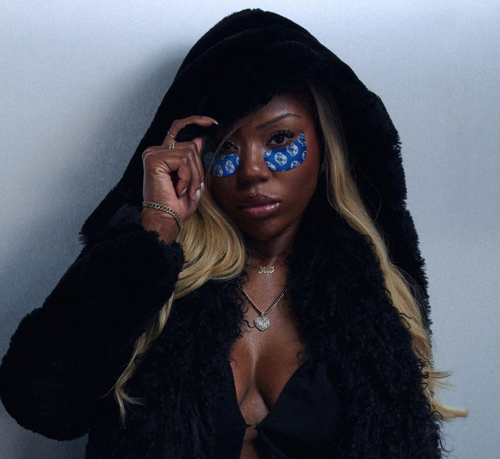 Brightening undereye patches from Topicals worn by a woman in a black hooded coat and gold jewelry, posing directly at the camera.